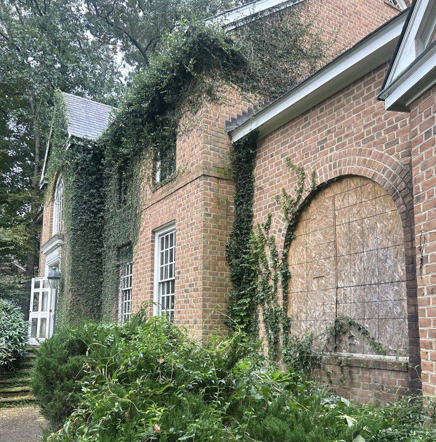 A brick house covered in ivy, with a boarded-up arched window and an overgrown front yard